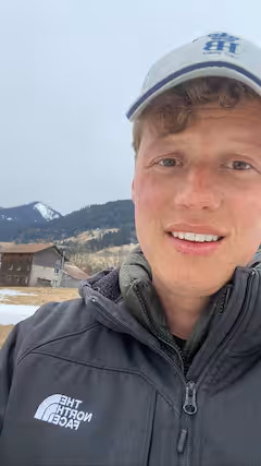 Jeune homme portant une veste North Face grise et une casquette blanche, en extérieur, avec des montagnes enneigées et des chalets en arrière-plan.